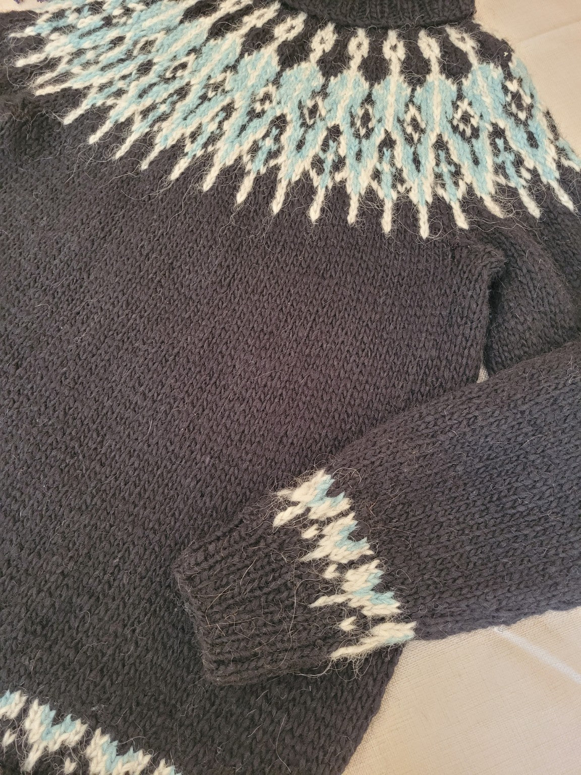 Detailaufnahme der handgestrickten Rundpasse des Islandpullovers Tusseladd aus Álafosslopi in Schwarz mit eisblauem und naturweißem Fair-Isle-Muster.