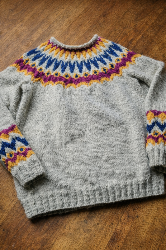 Handgestrickter Islandpullover „Loimüneule“ aus 100 % Léttlopi-Wolle, auf einem Holztisch liegend, mit farbigem Rundpassenmuster in Blau, Gelb und Beere.