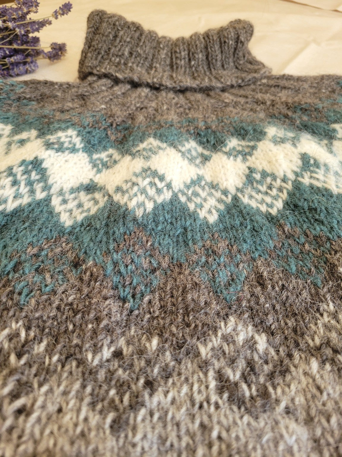 Detailaufnahme der handgestrickten Rundpasse eines Islandpullovers aus Lettlopi-Wolle in Grau, Petrol und Naturweiß mit traditionellem nordischem Muster.