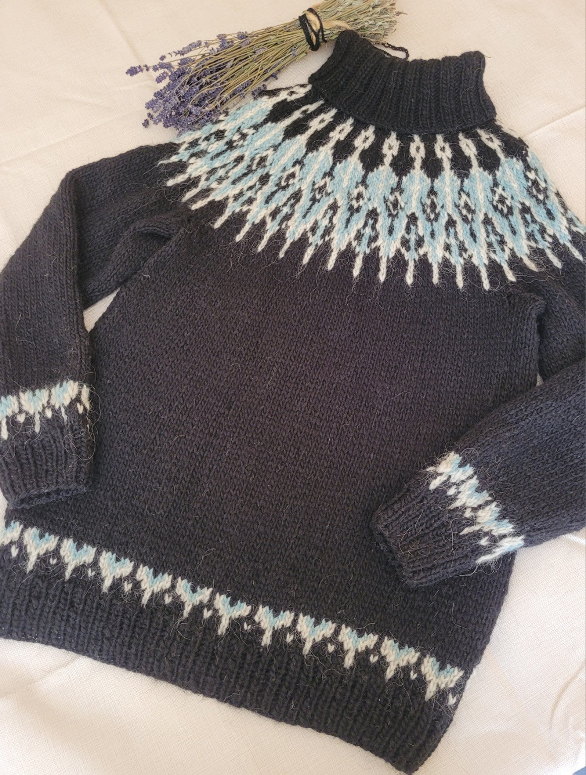 Handgestrickter Islandpullover Tusseladd aus Álafosslopi in Schwarz mit eisblauer und naturweißer Rundpasse, flach liegend fotografiert.