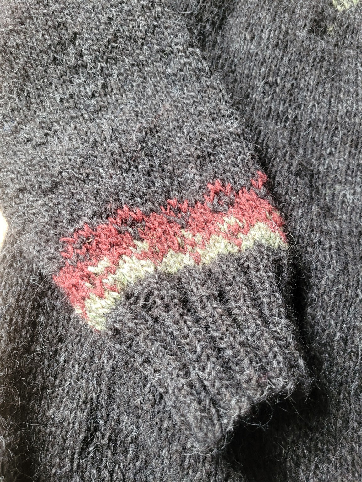 Nahaufnahme des Ärmelbündchens mit eingestricktem Muster in Rostrot und Salbei auf anthrazitfarbener Lopi-Wolle.