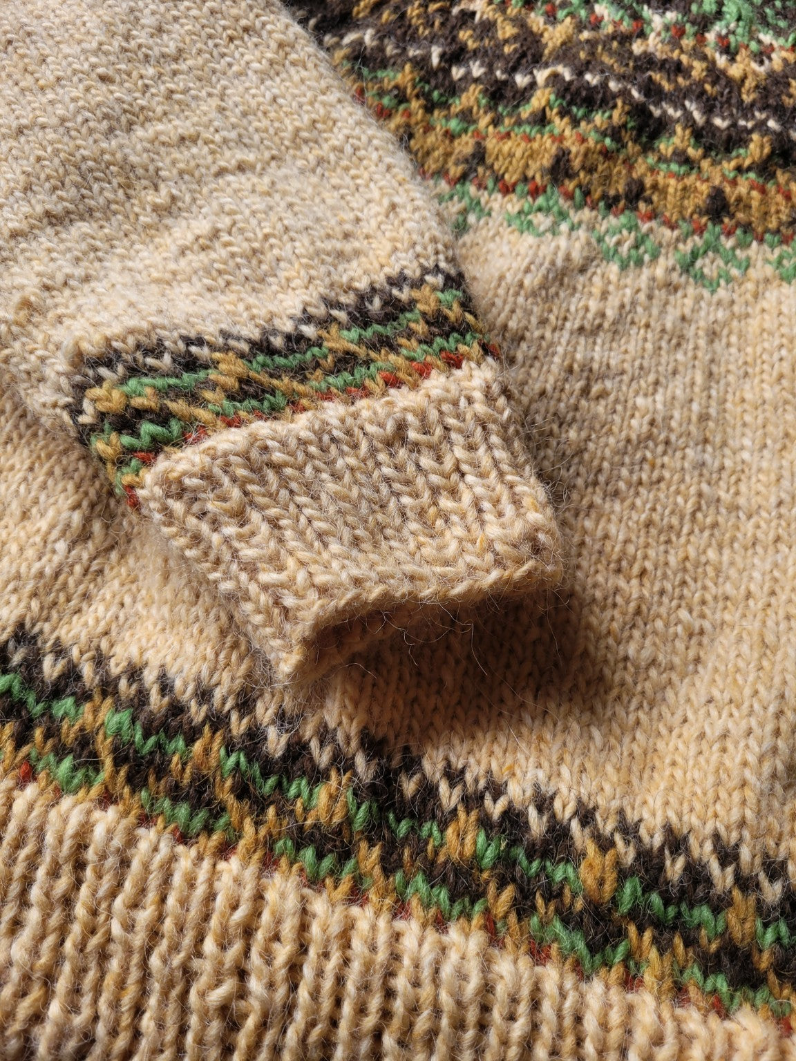 Detailaufnahme des Ärmelbündchens vom handgestrickten Islandpullover Alasuq mit feinem Farbverlauf in Grün, Braun und Senf auf naturfarbener Wolle.