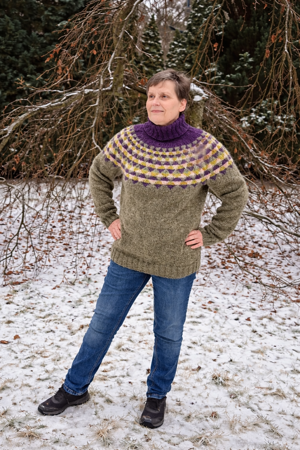 frau traegt handgestrickten islandpullover „bjoerk“ aus islaendischer lettlopi im winterlichen garten, rundpasse mit nordischem muster in violett, gruen und creme, stehkragen, alltagsnahes tragebild