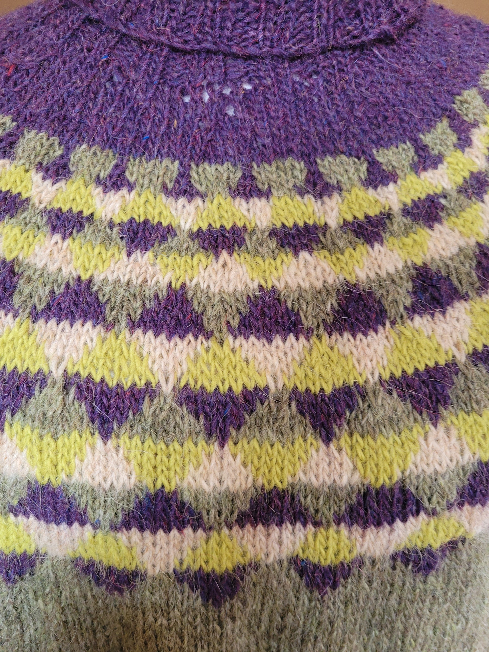 detailaufnahme des handgestrickten rundpassenmusters vom islandpullover „bjoerk“, geometrisches nordisches muster aus islaendischer lettlopi in violett, gruen und creme